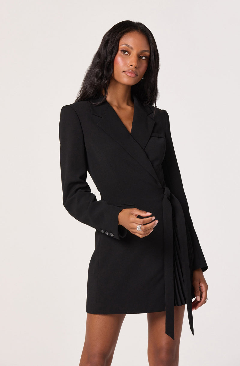 A woman is wearing a black blazer dress with a tailored fit. The dress features a notched lapel collar, long sleeves with buttons on the cuffs, and a waist belt tied in a bow. The hemline is above the knee.