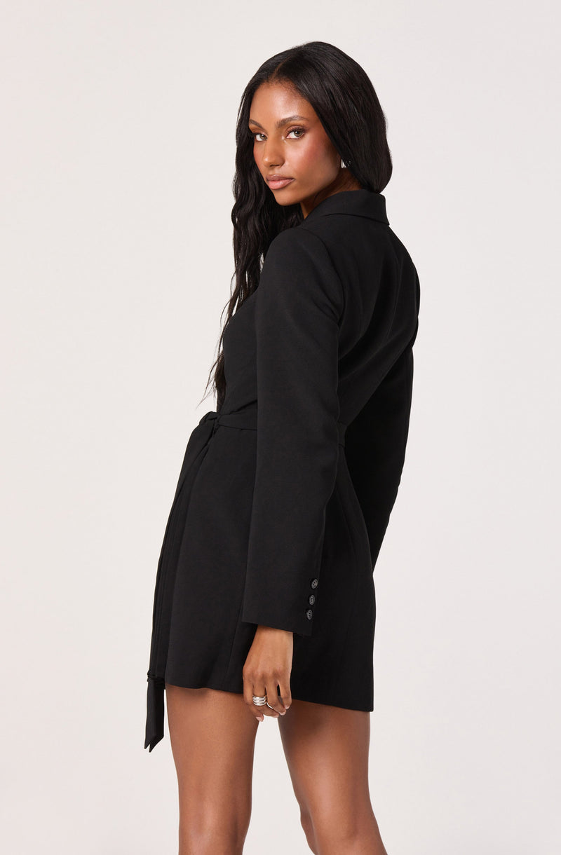 A woman is wearing a black blazer-style dress with long sleeves and a notched collar. The dress features a belt tied at the waist and button details on the sleeve cuff.