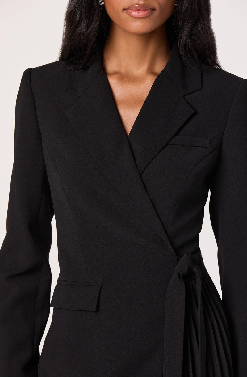 A woman is wearing a fitted black blazer with a structured lapel and shoulder pads. The blazer features a small pocket on the left side of the chest and a flap pocket on the right side near the waist. It also has a matching black fabric belt tied at the waist, creating a tailored silhouette.