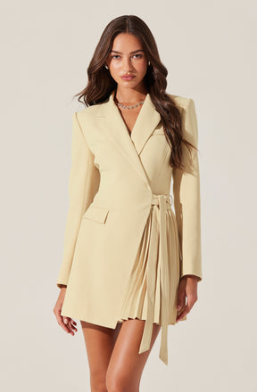 A woman is wearing a cream-colored blazer dress with padded shoulders, a notched lapel collar, long sleeves, and a single button closure at the waist. The dress features a pleated skirt detail on one side, with a fabric belt tied at the waist.