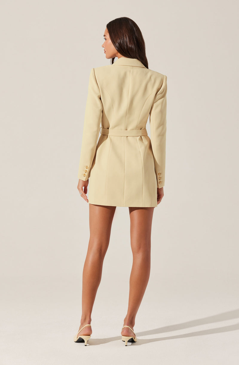 A woman is wearing a long beige blazer with a belt at the waist, featuring structured shoulders and button details on the sleeves. She is also wearing cream-colored high-heeled shoes with ankle straps.