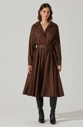 A woman is wearing a long brown shirt dress with a collar, button-down front, and long sleeves with cuffs. The dress features a matching belt at the waist, creating a gathered effect, and the skirt has pleats that give it a full, flowing silhouette. She is pairing the dress with knee-high black boots.