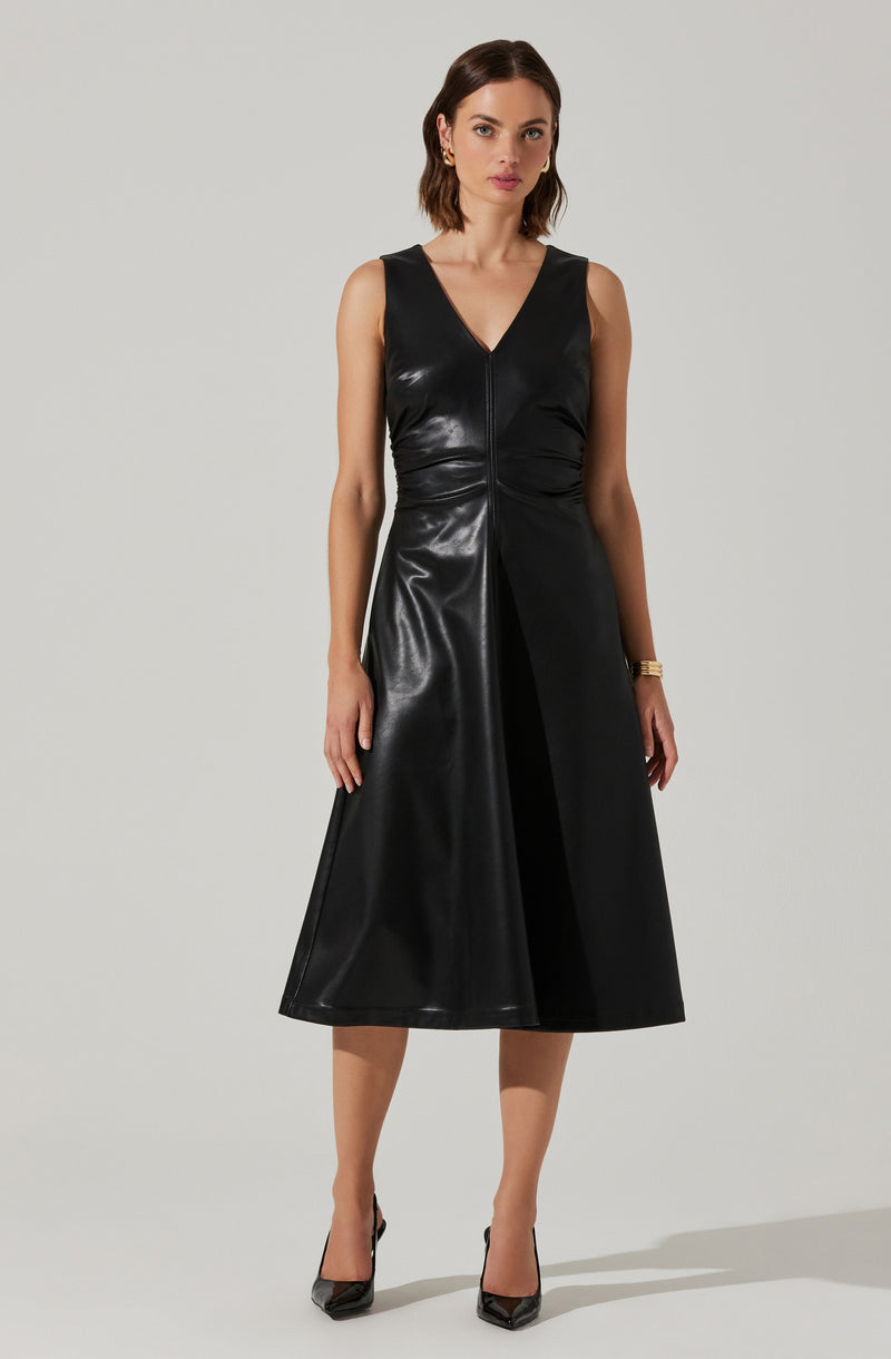 A woman is wearing a sleeveless, black dress made of a shiny, leather-like material. The dress has a V-neckline and a front zipper that runs from the neckline to the hem. It features a fitted bodice with subtle ruching at the waist and a full-length, A-line skirt that falls below the knees. She is accessorized with small earrings, a watch on her left wrist, and black pointed-toe high heels.