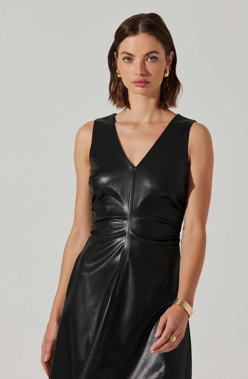 A woman is wearing a sleeveless black leather dress with a V-neckline and a front zipper running from the neckline to the hem. She has shoulder-length brown hair, gold earrings, and a gold bracelet on her left wrist.