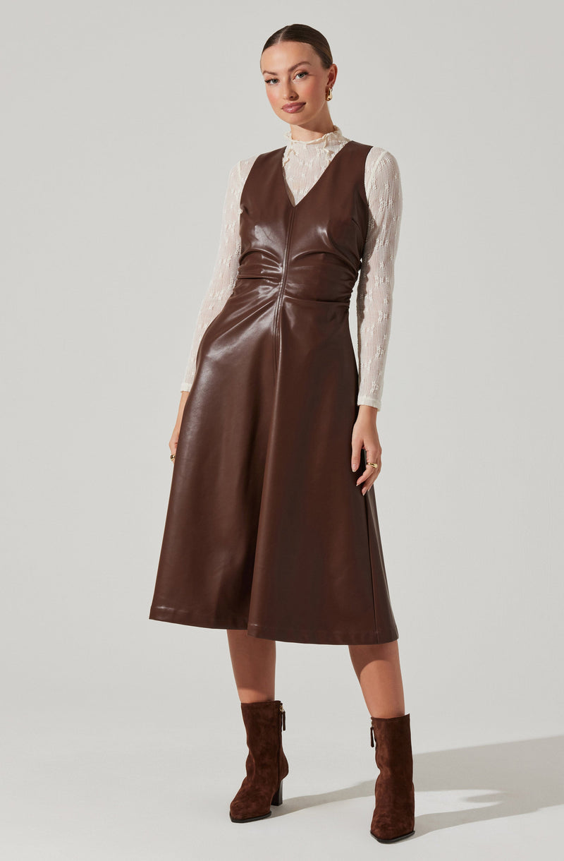 A woman is wearing a sleeveless brown leather dress with a deep V-neckline over a long-sleeved white lace blouse. She has paired the outfit with brown ankle boots that have a block heel.