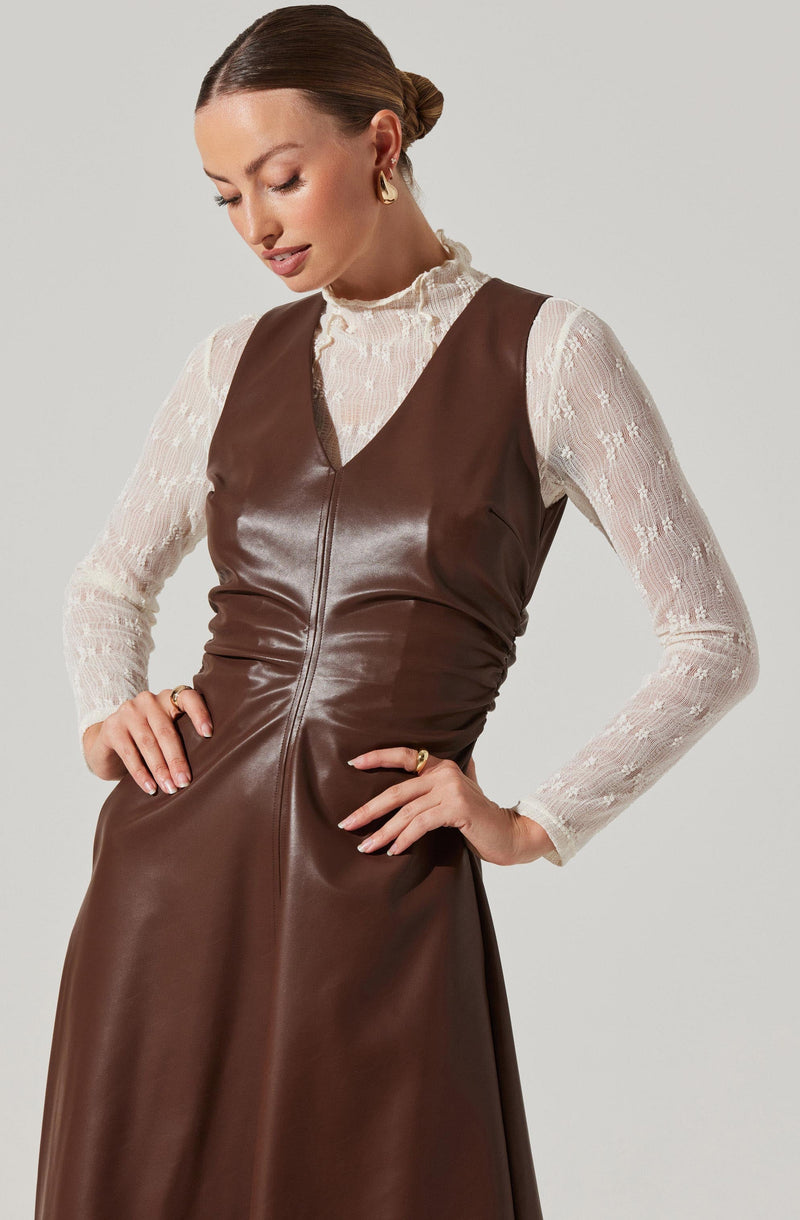A woman is wearing a sleeveless, brown leather dress with a high neckline. Underneath, she has on a long-sleeved, white lace top with a ruffled high collar and delicate floral patterns.