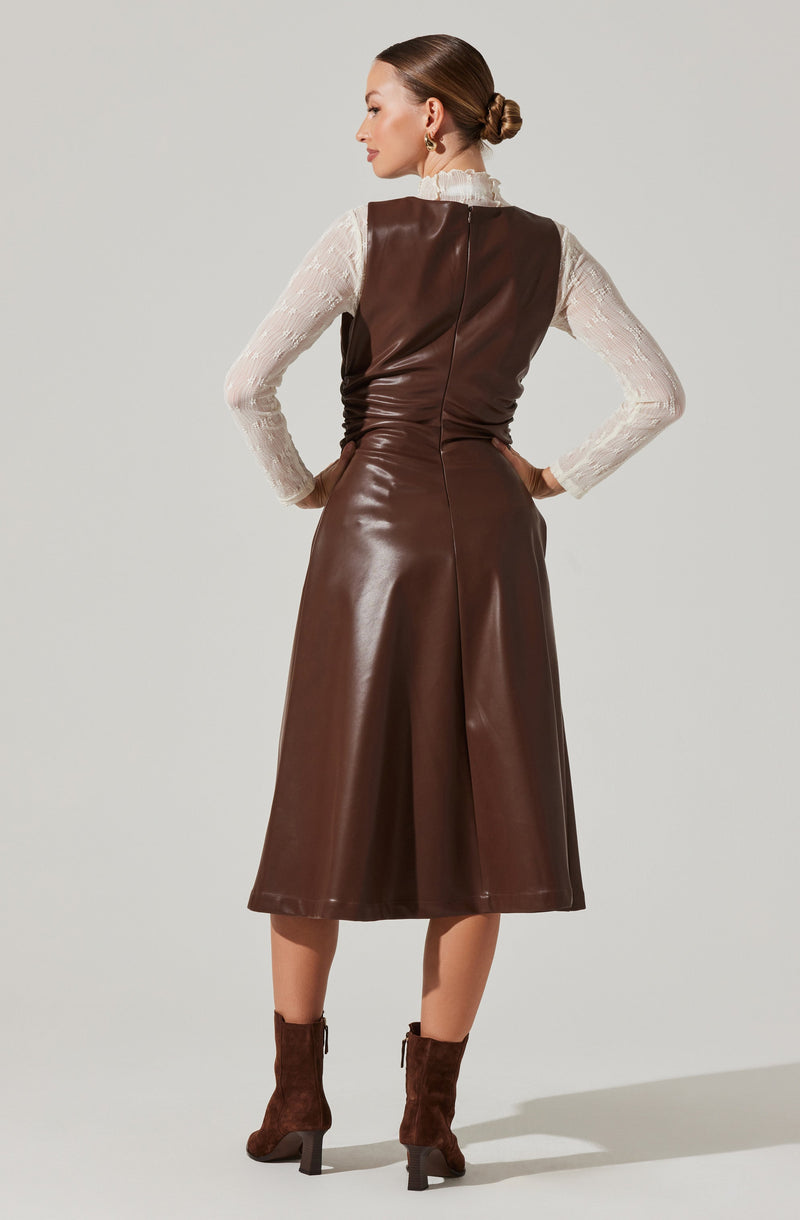 A woman is wearing a long, sleeveless, brown leather dress with a center back zipper. She has on a long-sleeved, cream-colored lace blouse with a ruffled high neckline underneath the dress. She pairs the outfit with brown suede ankle boots that have high block heels.