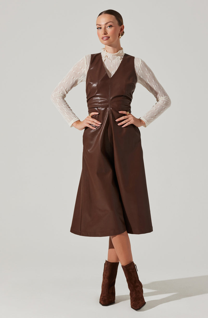A woman is wearing a sleeveless, brown leather dress that reaches mid-calf length, with a deep V neckline. Underneath, she has on a long-sleeved, white lace blouse with a high ruffled neckline. She pairs the outfit with brown suede ankle boots.