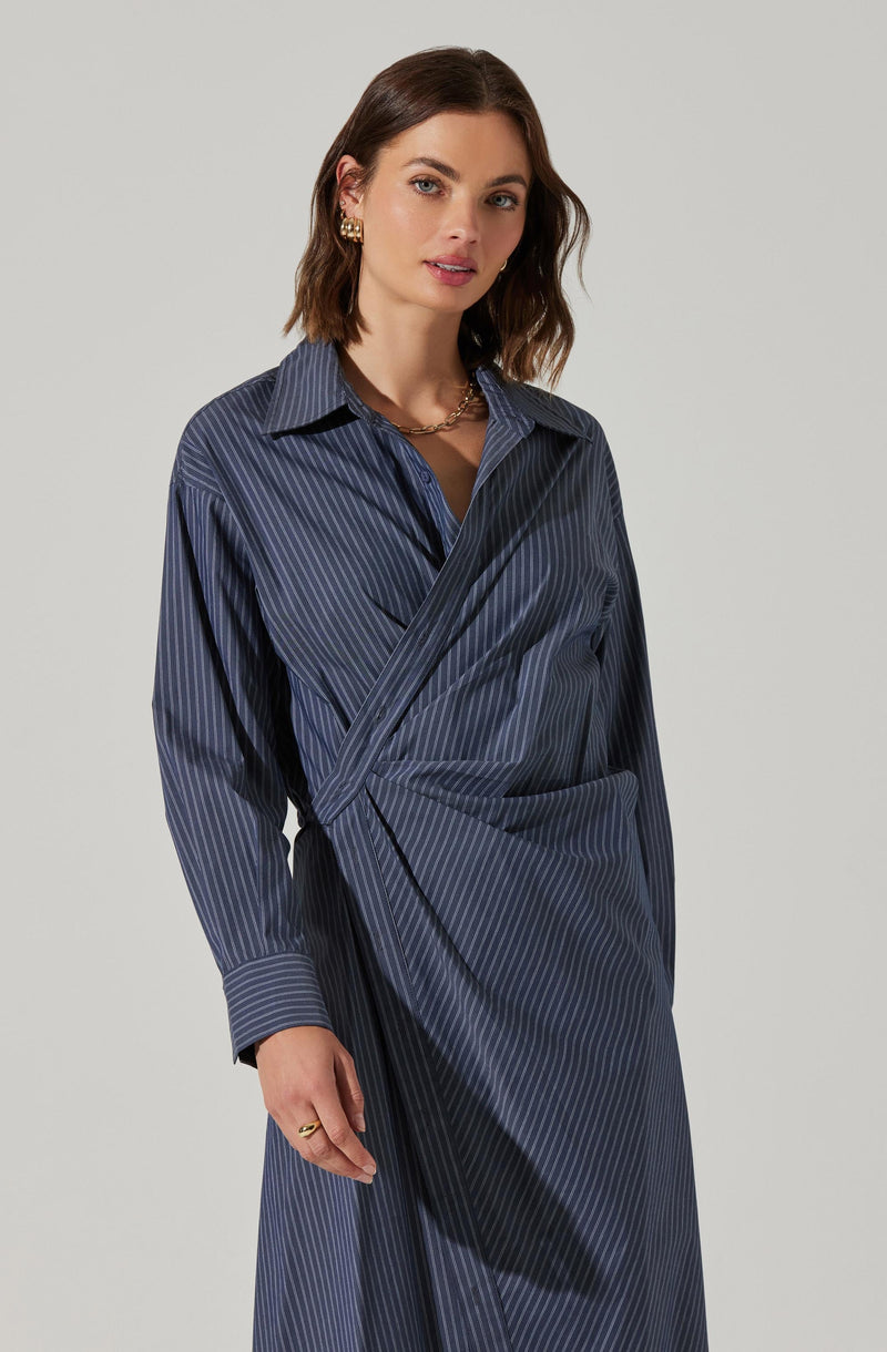 A woman is wearing a long-sleeved, dark blue shirt with light blue vertical pinstripes. Over the shirt, she has draped a matching fabric wrap that creates a layered look. The shirt features a classic collar and button-down front.