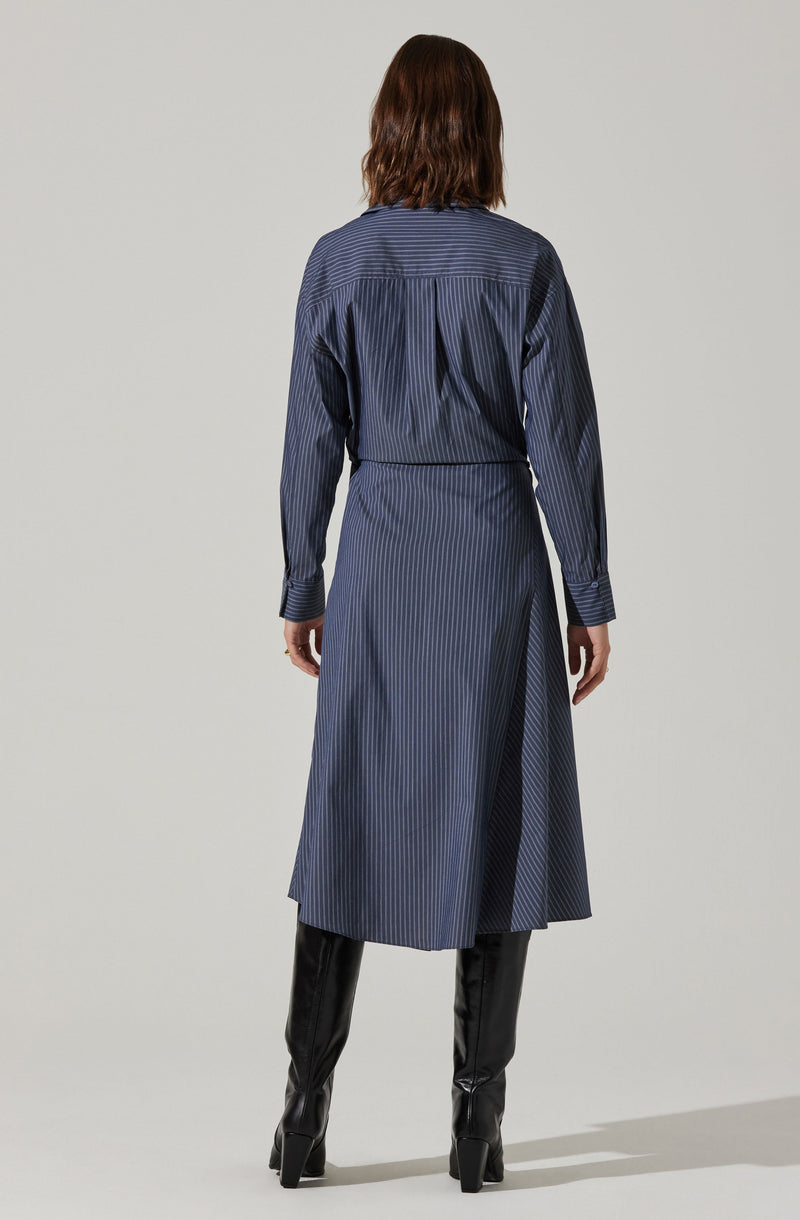 A woman is wearing a long, dark blue dress with vertical pinstripes. The dress has long sleeves, a high collar, and pleats on the back. She is also wearing tall, black knee-high boots with a chunky heel. The image shows her from the back, with her hands relaxed by her sides.