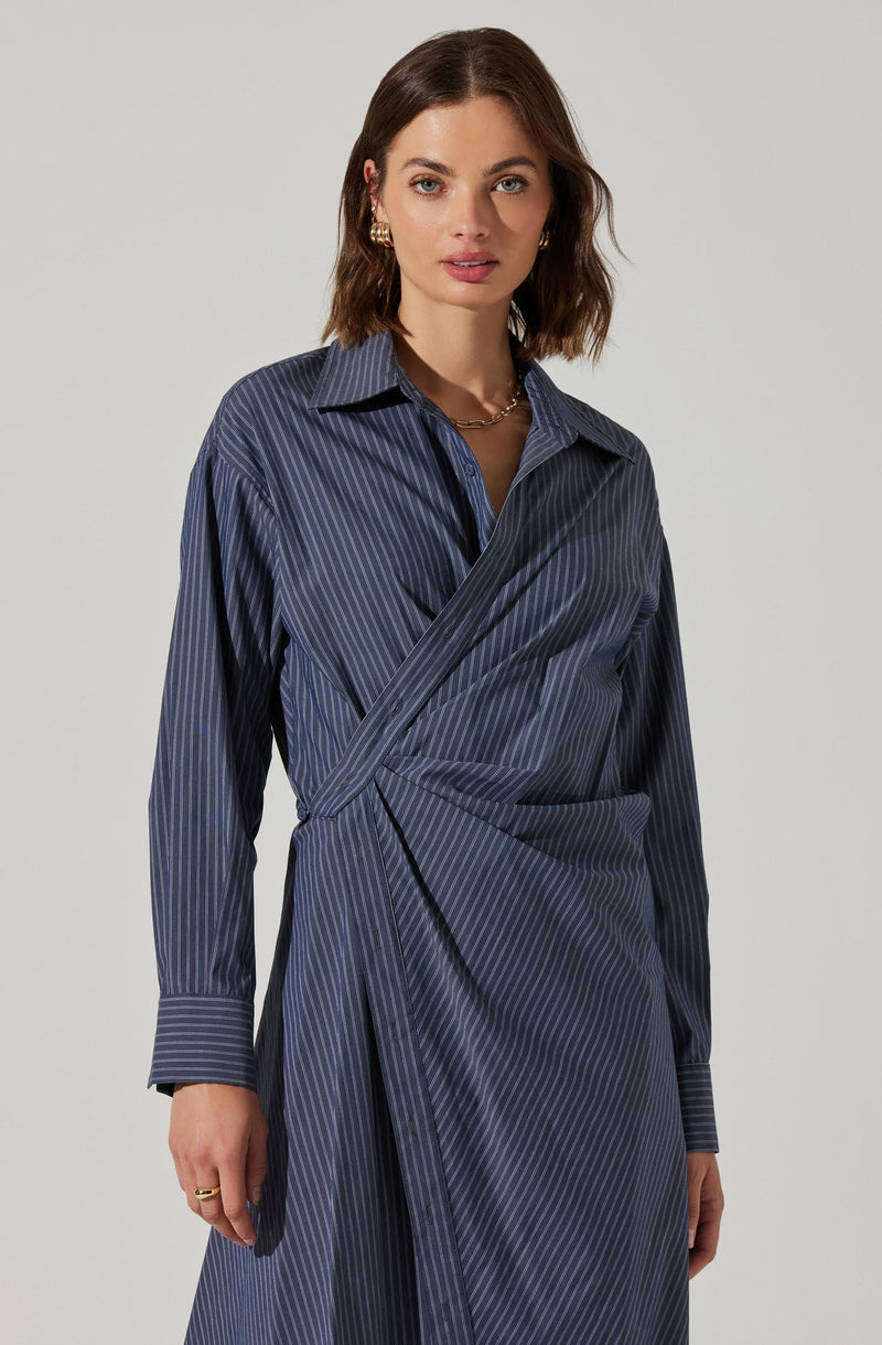 A woman is wearing a navy blue dress with thin, light-colored vertical pinstripes. The dress features a collared neckline, long sleeves with cuffs, and a wrap design that creates an asymmetrical, layered look at the waist.