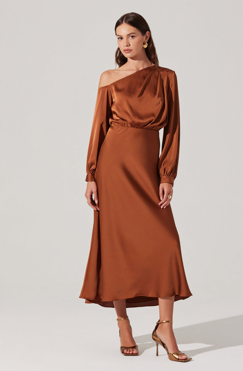 A woman is wearing a satin dress in a warm, copper-brown color. The dress features a one-shoulder neckline with an off-the-shoulder sleeve on one side and a long sleeve with elastic at the wrist on the other. It is fitted at the waist and flows into a long, slightly flared skirt that reaches the ankles. She completes the outfit with gold high-heeled sandals and gold earrings.