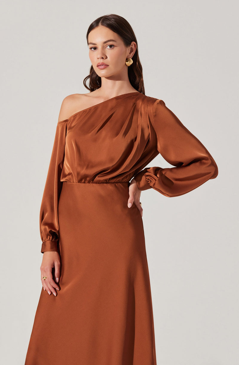 A woman is wearing an elegant, one-shoulder, satin-brocade dress in a rich, warm copper color. The dress features a draped bodice with gathered fabric at the shoulder and a loose fit at the waist, with the long sleeve on the left side gathered at the wrist. The skirt is straight and extends to the floor, creating a sleek silhouette. She accessorizes with large, gold, textured earrings.