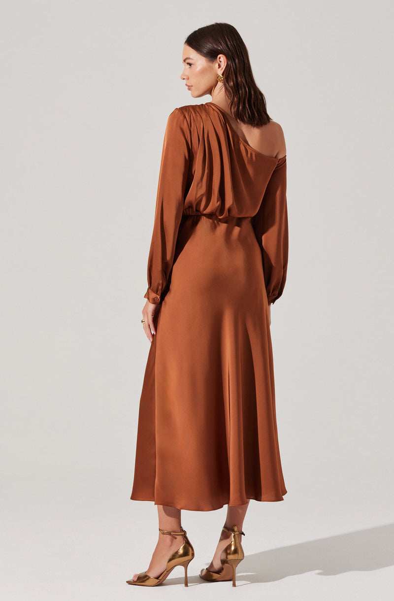 A woman is wearing a long, elegant, bronze-colored dress with long, puffed sleeves gathered at the wrists. The dress has a gathered neckline that drapes off one shoulder, creating a stylish asymmetrical look. The waist is cinched, and the skirt flows down to mid-calf length with a smooth, shiny satin finish. She pairs the outfit with gold high-heeled sandals and gold earrings.