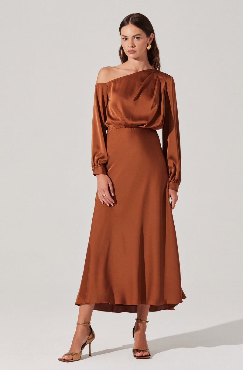 A woman is wearing a long, elegant, satin dress in a rich bronze or copper color. The dress features an off-the-shoulder neckline with long sleeves that have elasticized cuffs. It has a fitted waist and a flowing, ankle-length skirt. She is accessorizing with gold hoop earrings and open-toe high-heeled sandals.