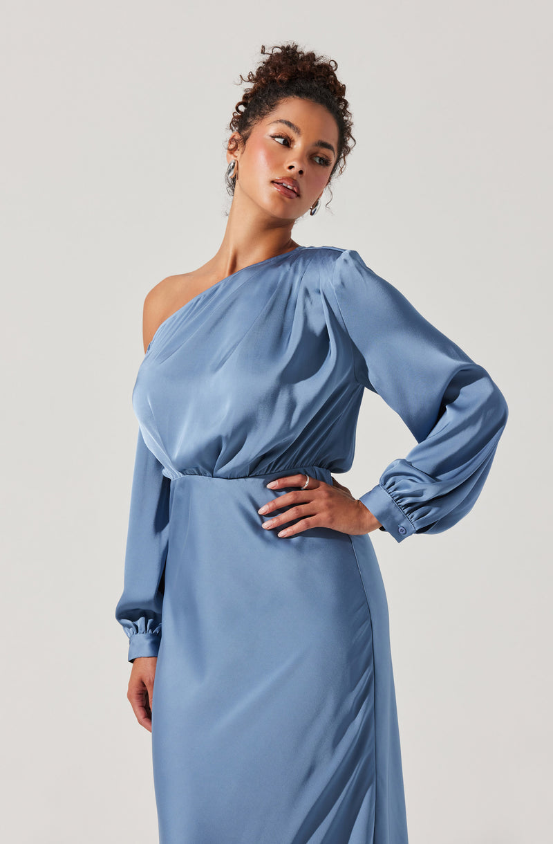 A woman is wearing a light blue satin dress with a one-shoulder design. The dress has long, puffed sleeves that gather at the wrists with buttoned cuffs. The waistline is defined with a fitted elastic band, and the skirt falls straight with a smooth, shiny finish.