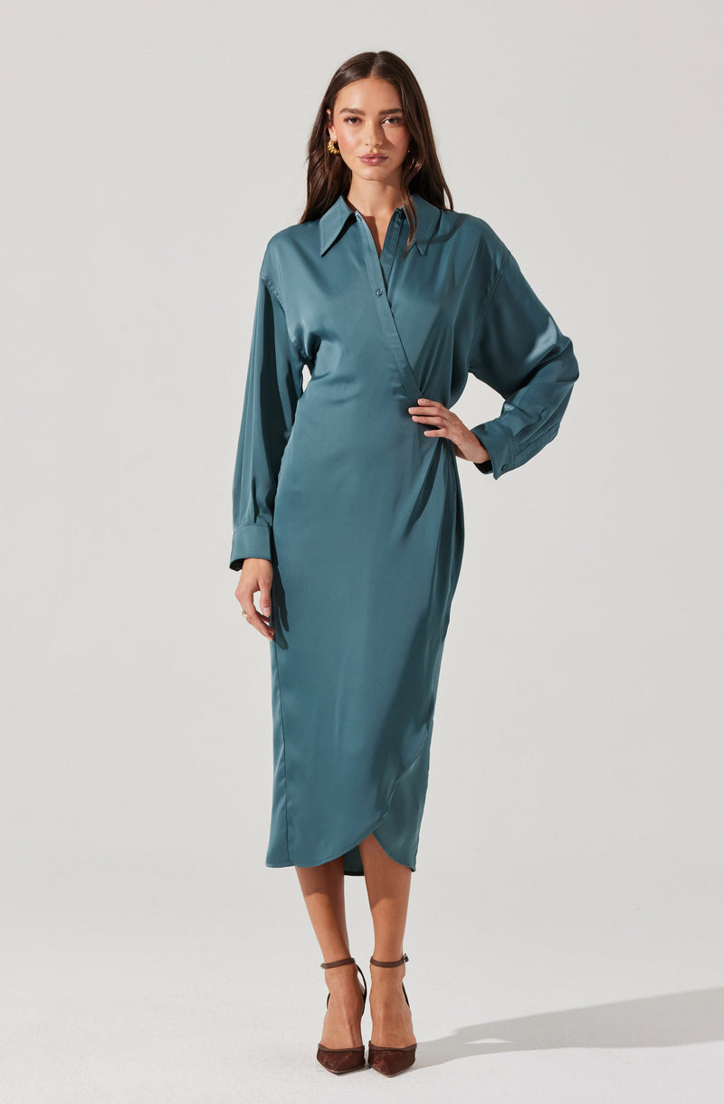 A woman is wearing a long, silky, teal-colored shirt dress with a pointed collar and button-down front. The dress has long sleeves with buttoned cuffs and a side slit at the hem. She is accessorizing with gold earrings and brown high-heeled shoes.