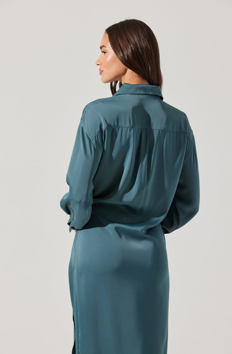 A woman is wearing a long-sleeved, teal satin blouse with a pointed collar, and a matching teal satin skirt. The blouse has a loose fit with gathered fabric at the back yoke. The skirt is smooth and shiny, with a side slit visible. The woman's hair is straight and brown, and she is standing against a plain, light-colored background.
