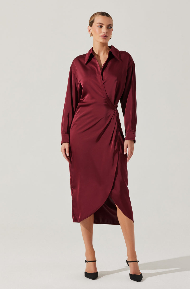 A woman is wearing a long-sleeved, deep burgundy satin dress with a classic collar and a wrap-style skirt that features an asymmetrical hemline with a slit. She is also wearing black high-heeled shoes with ankle straps.