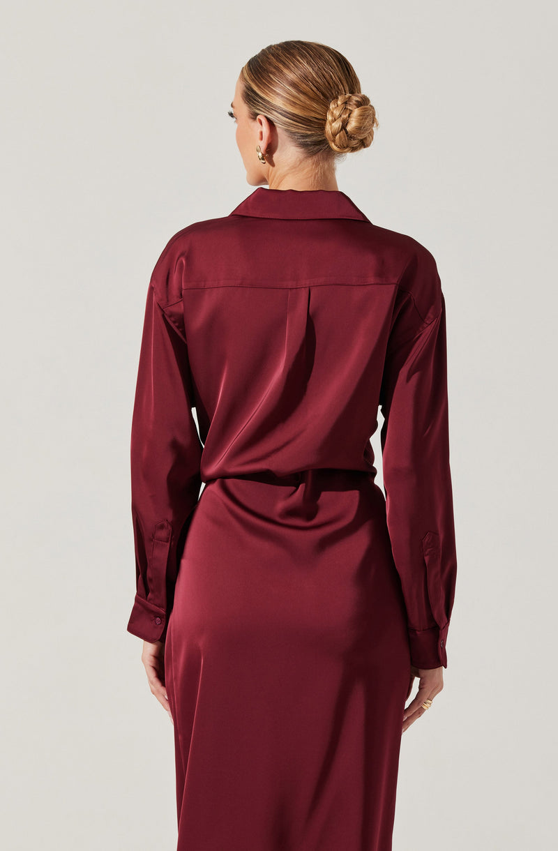 A woman is wearing a long-sleeved satin blouse and a matching satin skirt in a deep red or burgundy color. The blouse has a collar and buttoned cuffs, with a loose fit at the back. The skirt is fitted at the waist and flows down smoothly. The woman's hair is styled in a twisted bun, and she is accessorized with earrings and rings.