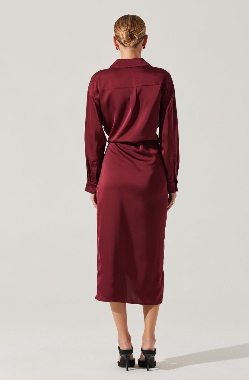 A woman is wearing a long, burgundy satin dress with a matching satin shirt tucked into the skirt. She has her hair styled in an elegant updo and is wearing black high-heeled shoes.