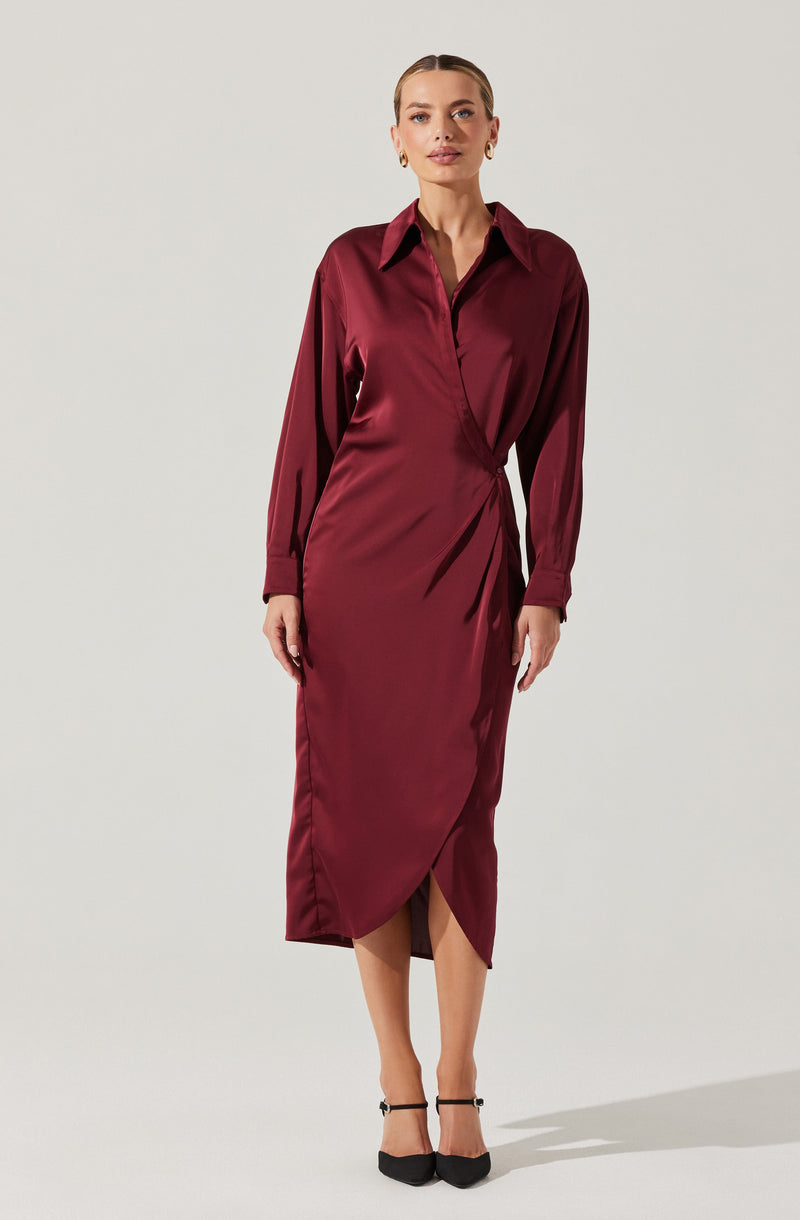 A woman is wearing a long, satin-like dress in a deep burgundy color. The dress has a collared neckline with a button-down front. It features long sleeves with cuffs, and the waist is slightly gathered on one side, creating a draped effect. The dress extends to mid-calf length with a front slit that reveals a portion of her legs. She is also wearing black, pointed-toe high heels with ankle straps.