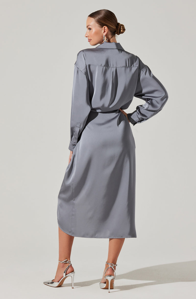 A woman is wearing an elegant, long, silver satin dress with a collared top and long sleeves that are gathered at the cuffs. The dress has a cinched waist and a flowing skirt that extends to her calves. She is also wearing high-heeled sandals with ankle straps, and her hair is styled in a sleek, low bun.