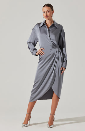 A woman is wearing a long, shiny, silver-gray satin dress with a collared neckline and long, slightly oversized sleeves. The dress features a draped design with pleats on one side, creating an asymmetrical, wrapped appearance. She is also wearing pointed silver high-heeled shoes with ankle straps.