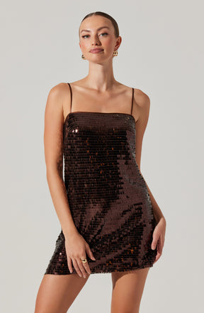 A woman is wearing a shiny, dark brown, sequined mini dress with thin spaghetti straps. The dress fits snugly and has a straight neckline. She is accessorized with gold hoop earrings and a ring on her right hand.