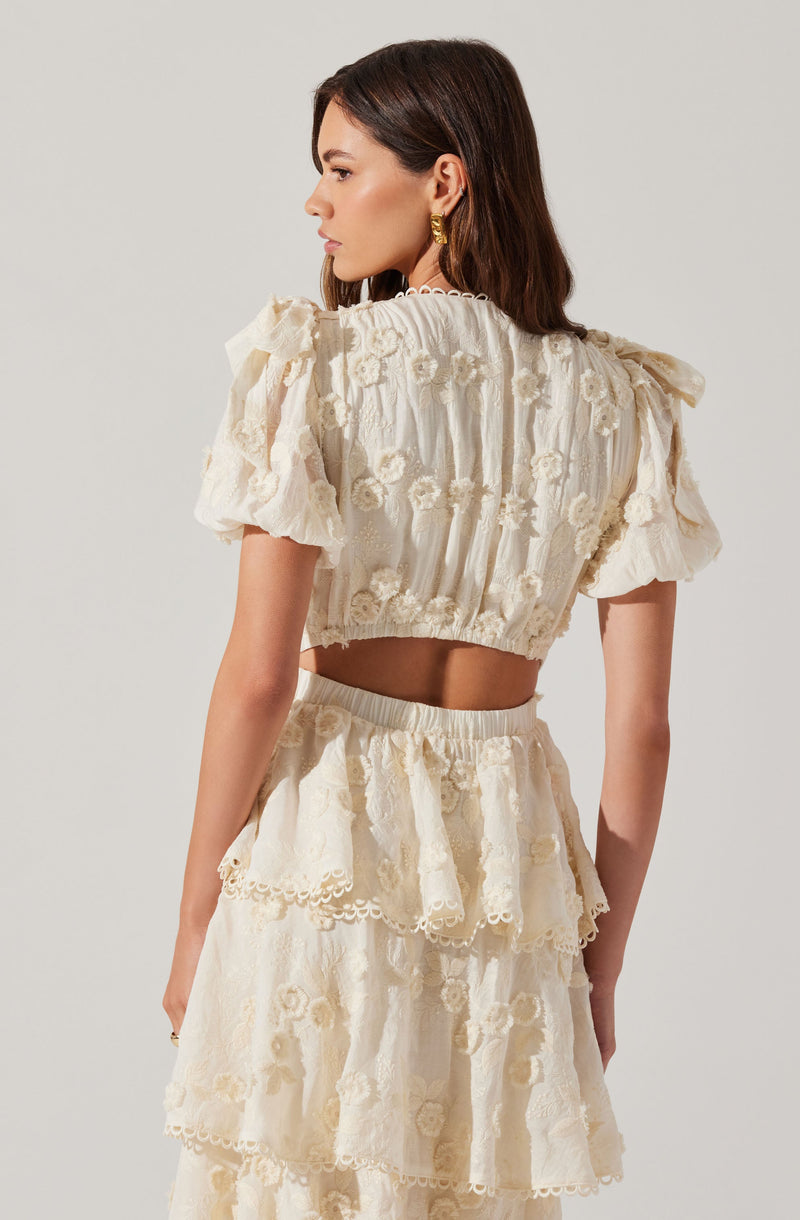 A woman is wearing a matching cream-colored two-piece outfit consisting of a cropped top with puffed short sleeves and a high-waisted skirt. Both pieces feature intricate floral embroidery and lace detailing. The top has a loose fit with ruffled accents, and the skirt includes layered, ruffled tiers with decorative edges.