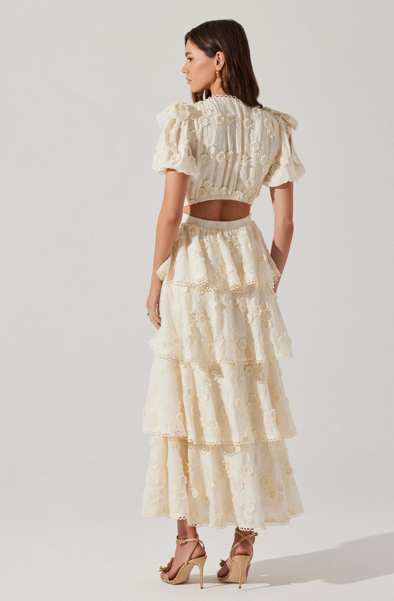 A woman is wearing an elegant, cream-colored two-piece outfit. The top is a short, cropped blouse with puffed sleeves and intricate floral embroidery. The skirt is high-waisted with multiple tiered layers, each adorned with delicate lace trim and embroidered floral patterns, creating a flowing, tiered silhouette. She is also wearing open-toe, ankle-strap high heels.