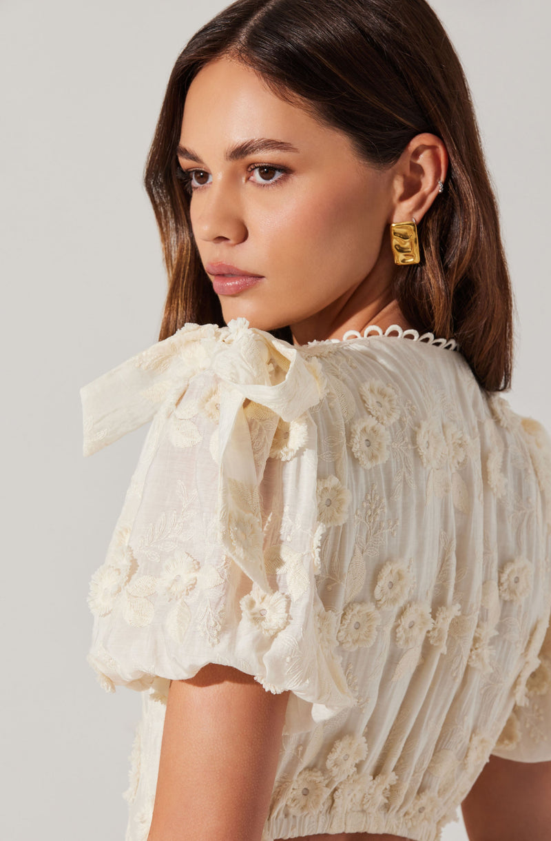 A woman is wearing a light-colored, short-sleeved blouse with floral embroidery. The blouse features a bow detail on the shoulder and a scalloped neckline. She has a gold rectangular earring and a small hoop earring in her ear.