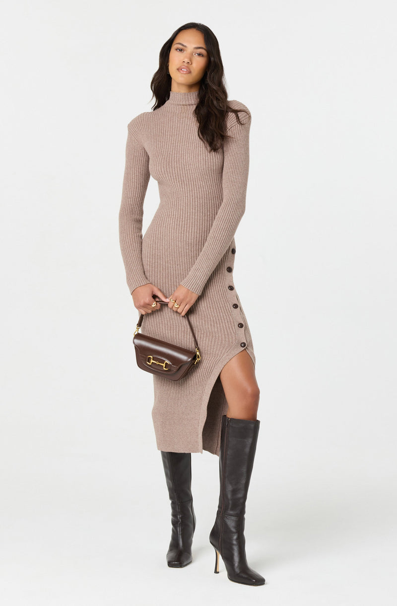 A woman is wearing a fitted, long-sleeve, ribbed-knit dress in a beige/taupe color with a high neckline. The dress features a side slit with buttons running down the length, exposing part of her leg. She is accessorizing with a small, dark brown handbag with gold hardware and is wearing black knee-high heeled boots.