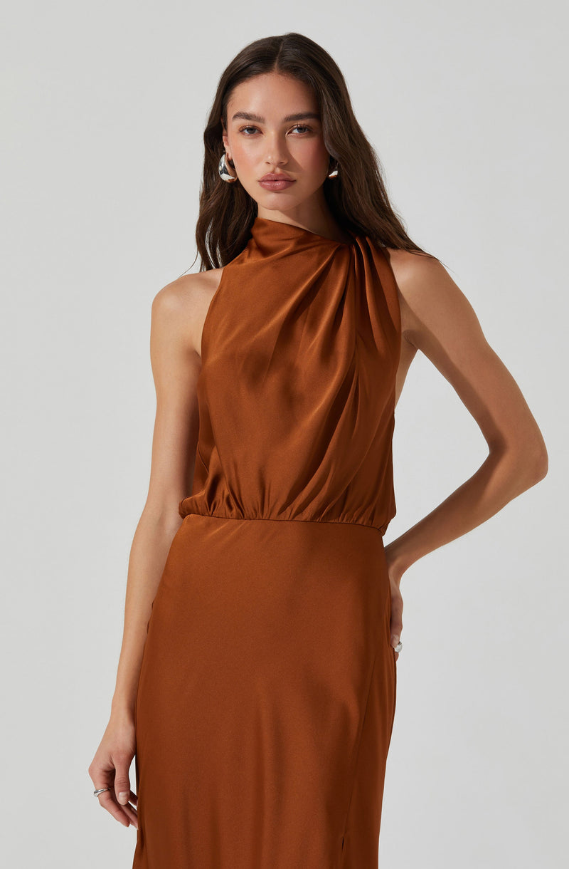 A woman is wearing a sleeveless, high-neck, satin dress in a rich, copper-brown color. The top of the dress features gathered fabric that drapes slightly across the chest, creating a soft, elegant look. The waistline is defined, and the skirt extends downward, maintaining a smooth and flowing appearance. She accessorizes with large, silver hoop earrings.