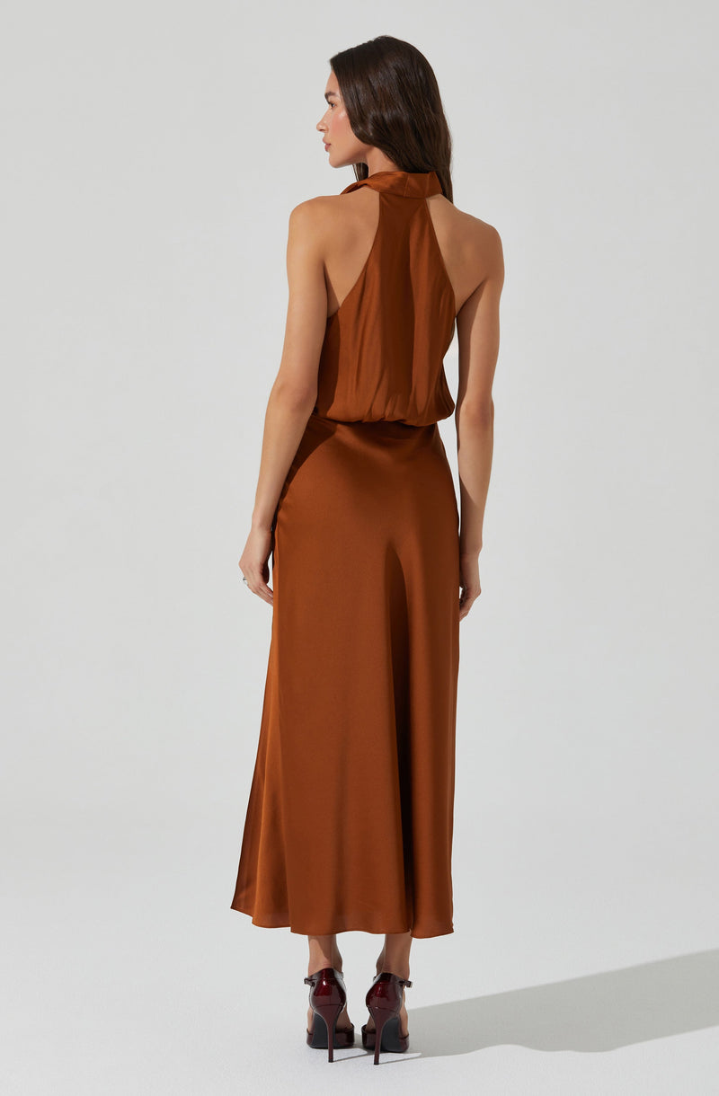 A woman is wearing a sleeveless, high-neck halter top made of a sheer, shiny, rust-colored fabric that drapes loosely. She pairs it with a long, flowing, satin-looking skirt in a matching rust hue that reaches her ankles. She is also wearing high-heeled shoes with a dark, glossy finish.