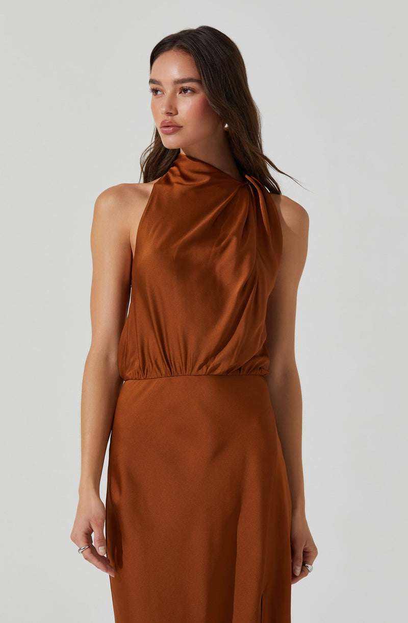 A woman is wearing a sleeveless, satin dress in a rich, burnt orange color. The dress features a high neckline with draped fabric that gathers at one shoulder, creating a flowing and elegant silhouette. The waist is fitted, and the fabric has a smooth, shiny finish.
