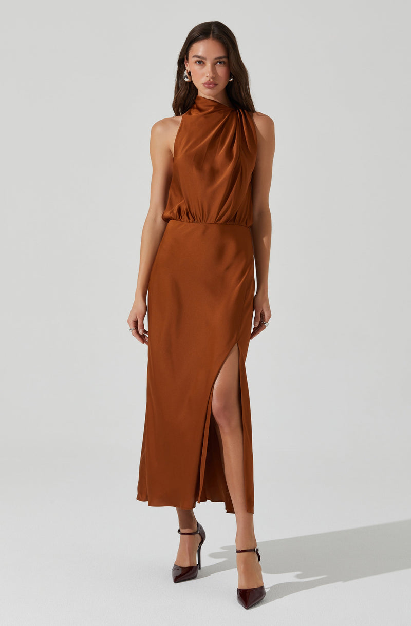A woman is wearing a sleeveless, high-neck satin dress in a rich, bronze-brown color. The top of the dress features soft draping, while the skirt is long and fitted with a side slit that reveals her leg. She pairs the dress with dark, pointed-toe heels and accessorizes with large earrings.