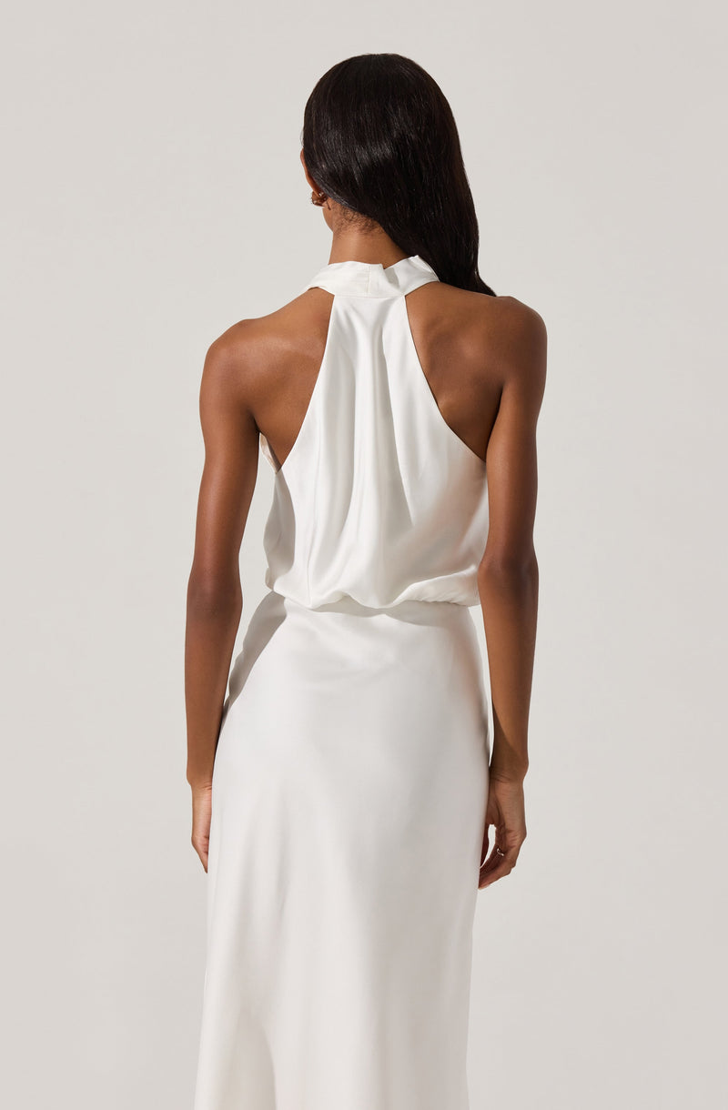 A woman is wearing a sleeveless, high-neck, white satin dress with a loose draped top and a fitted, floor-length skirt. The back of the dress features a gathered, flowing design.
