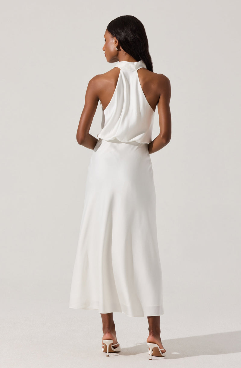 A woman is wearing a sleeveless, high-neck, white halter top with a loose, draped fit that ties at the back of her neck. She pairs it with wide-leg white pants that reach her ankles. She is also wearing white high-heeled sandals with thin straps. Her hair is long and black, and she is standing against a plain light-colored background.