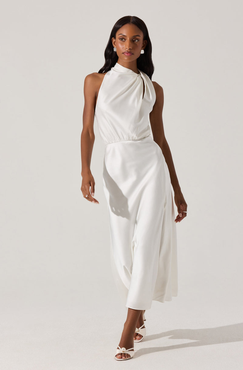 The woman is wearing a sleeveless, high-neck white jumpsuit with a gathered detail at the neckline and a fitted waist. The pants are wide-legged and flowy, reaching just above the ankles. She is also wearing white open-toe heels with ankle straps.
