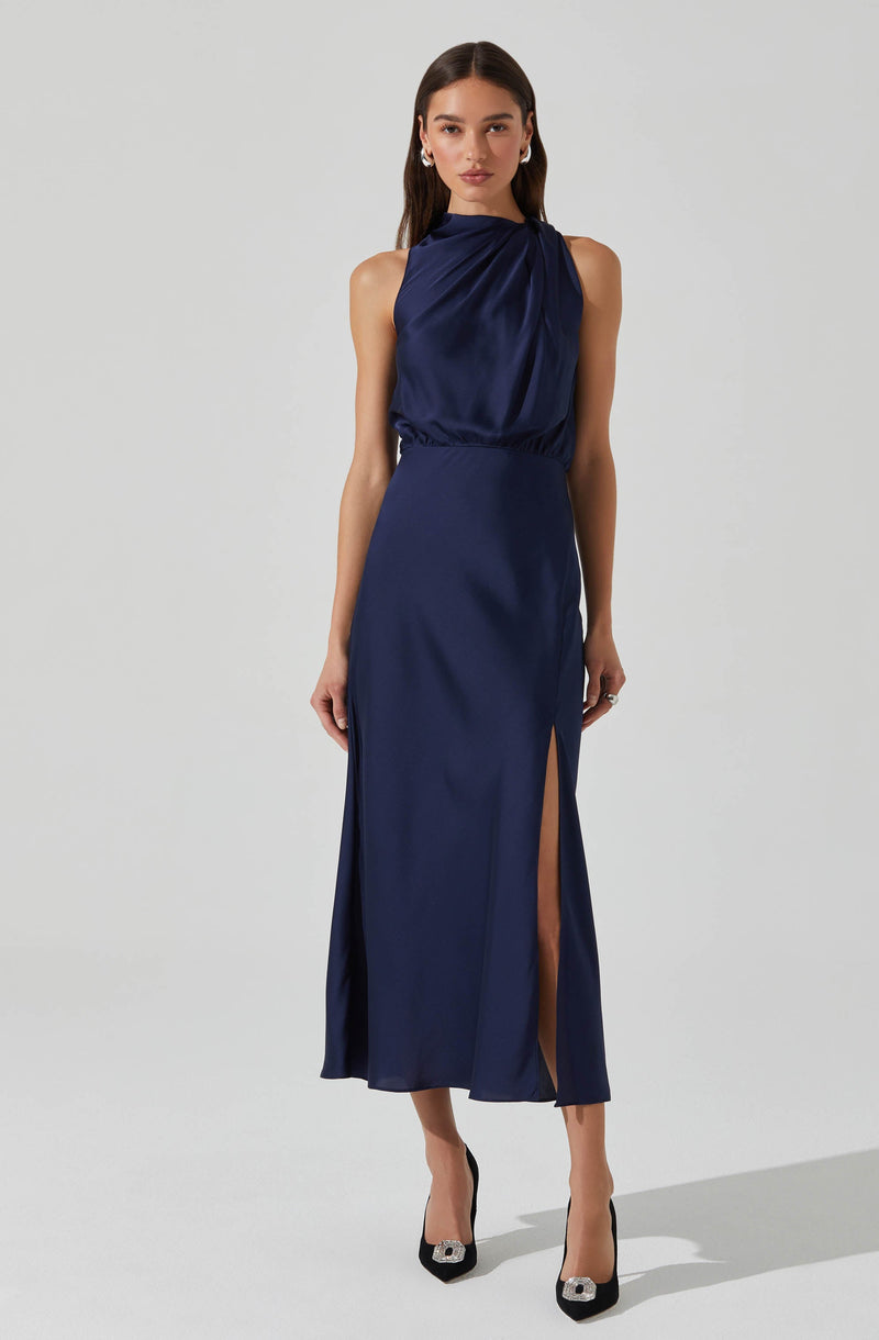 A woman is wearing a sleeveless, dark blue satin evening gown with a gathered, draped neckline and an elastic waistband. The dress has a high slit on the left side, revealing her leg. She is also wearing black high heels with decorative embellishments on the toes.