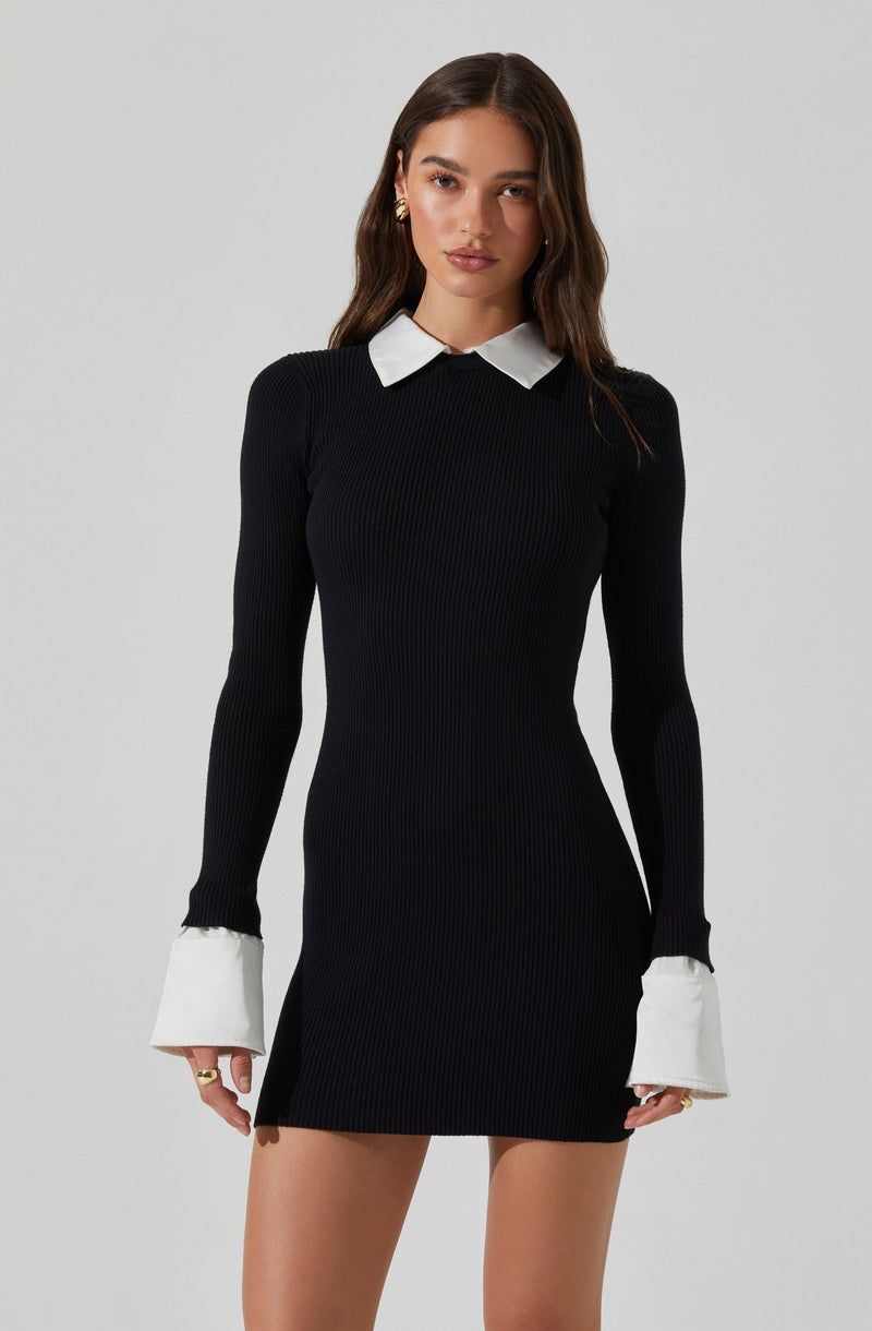 A woman is wearing a fitted, long-sleeve black ribbed knit dress with white contrasting collar and cuffs. The dress is short, ending above the knees, and the cuffs are designed with added white fabric that extends beyond the sleeves.