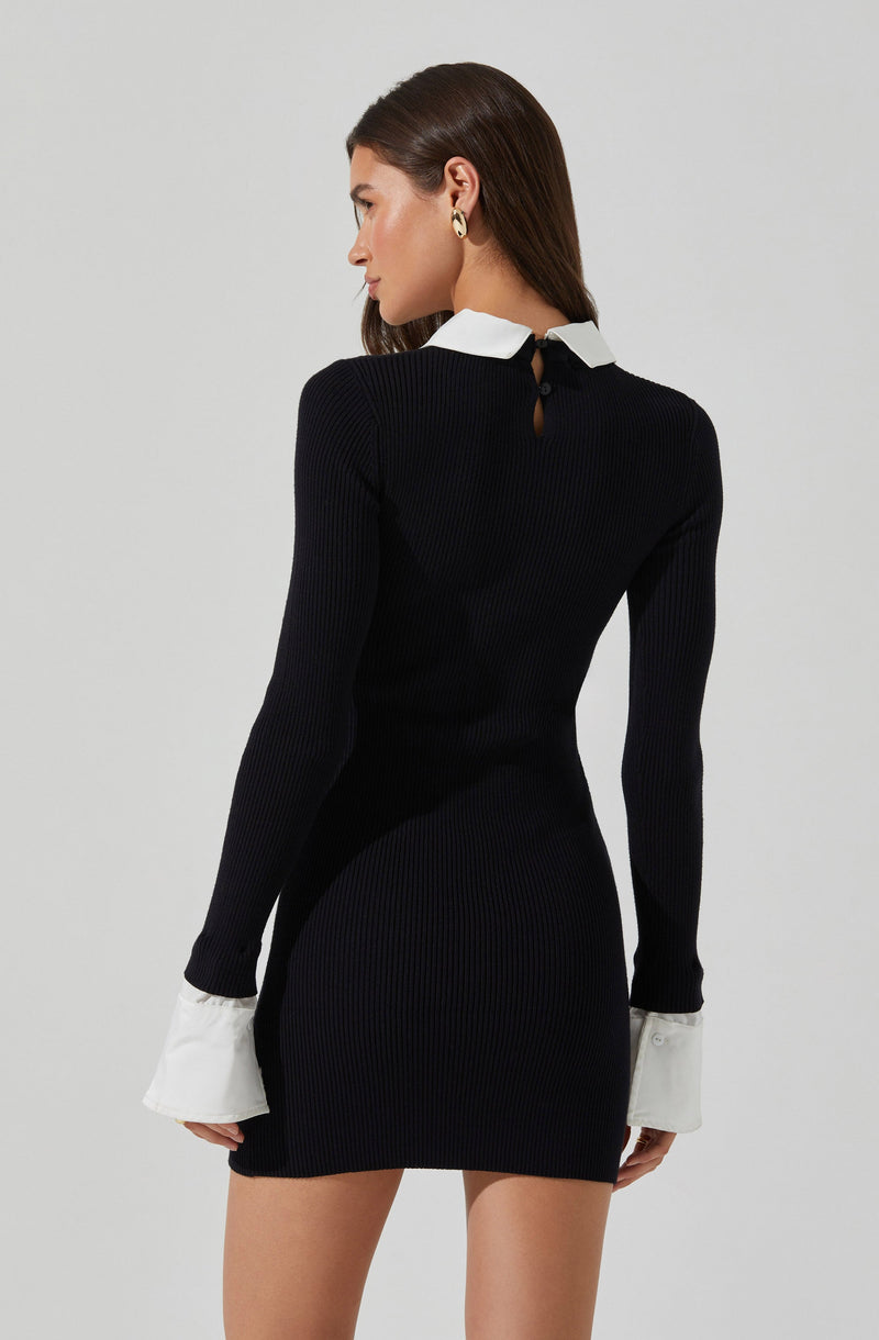 A woman is wearing a fitted, long-sleeved black ribbed knit dress with white cuffs at the sleeve ends and a contrasting white collar. The dress features a small keyhole opening at the back, fastened with a button.