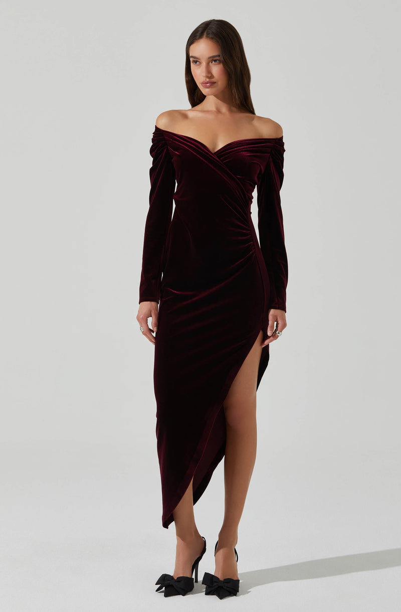 A woman is wearing an off-the-shoulder, long-sleeved dress made of deep burgundy velvet fabric. The dress features a sweetheart neckline with pleated detailing across the bust, a fitted waist, and a high slit on the left side that reveals her leg. She is also wearing black high heels with large bow accents on the toes.
