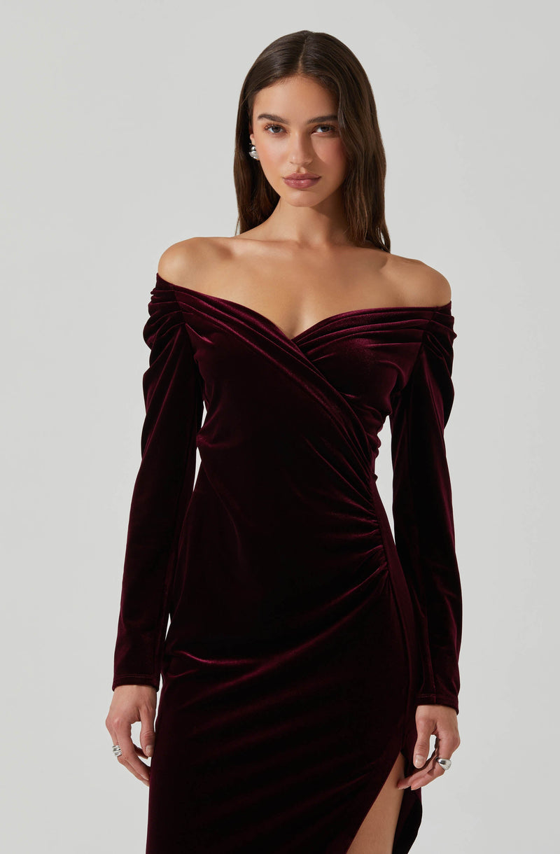 A woman is wearing a long, off-the-shoulder dress made of deep burgundy velvet fabric. The dress has long sleeves with gathered detailing at the shoulders and a fitted silhouette that accentuates her figure. There is a subtle slit on one side of the skirt.
