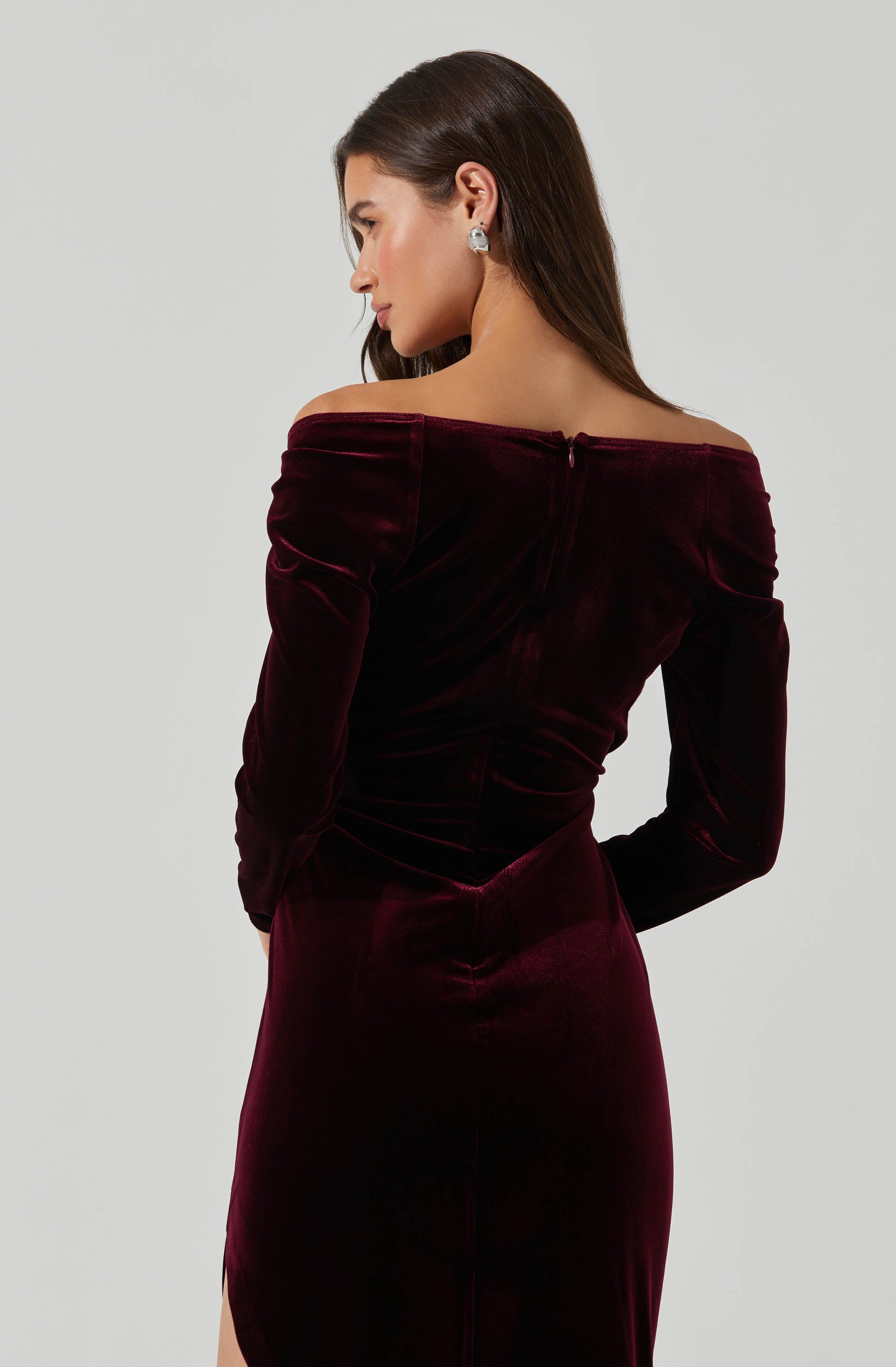 A woman is wearing an off-the-shoulder, long-sleeved dress made of dark red velvet fabric. The dress fits snugly and has a back zipper. She has long, dark brown hair and is wearing large, square-shaped earrings.