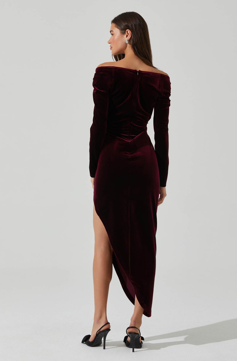A woman is wearing a long, off-the-shoulder velvet dress in a deep burgundy color. The dress has long sleeves and a fitted silhouette, with a slit on one side revealing her leg. She is also wearing black high-heeled sandals.