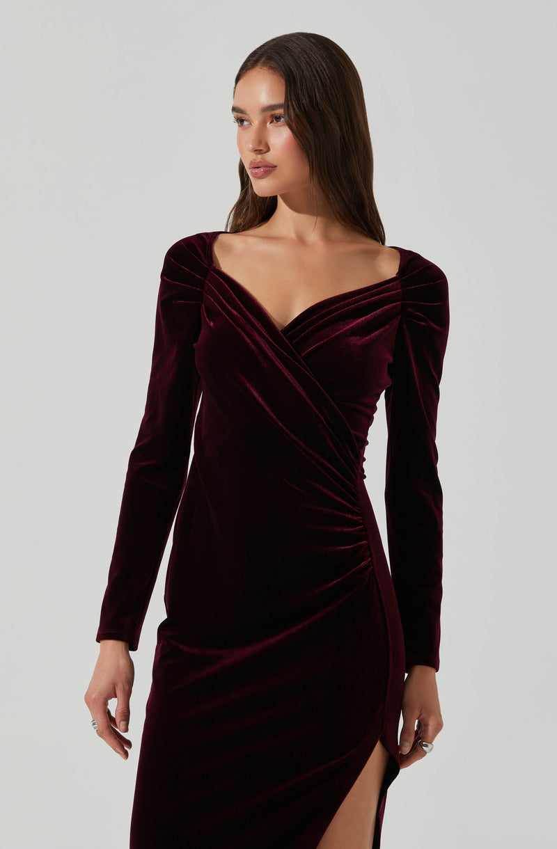 A woman is wearing a long, fitted, dark red velvet dress with long sleeves. The dress features a wrap-style bodice with a deep neckline and gathered detailing at the shoulders and waist, along with a high slit on one side.