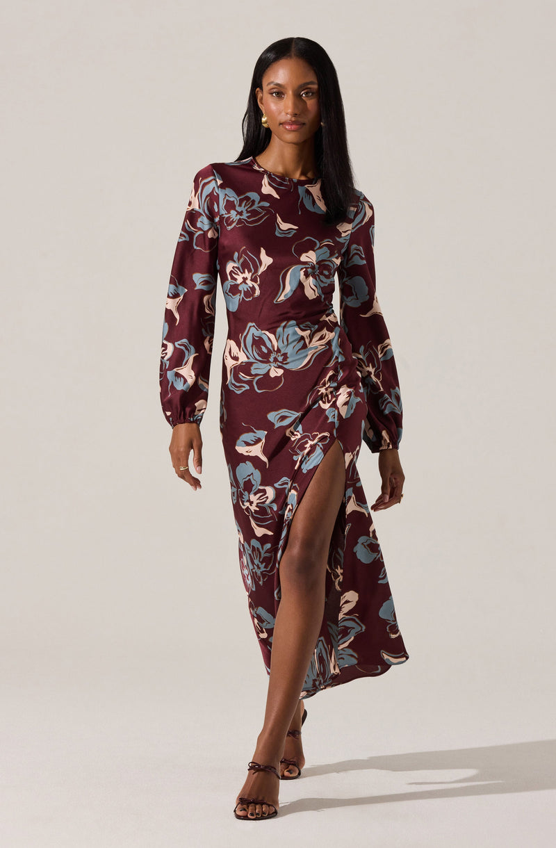 A woman is wearing a long-sleeved, midi-length dress with a high slit. The dress has a dark maroon background with large, abstract floral patterns in shades of blue, cream, and light pink. She is also wearing high-heeled sandals.
