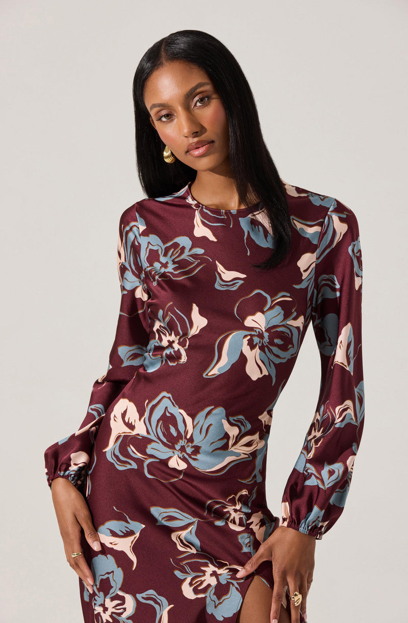 A woman is wearing a long-sleeved, satin dress with a floral pattern. The dress is primarily maroon with large, abstract flowers in shades of blue, cream, and brown. The fabric appears smooth and shiny, and the dress has a fitted silhouette with a slit on one side.
